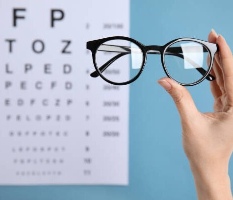 Woman holding glasses against eye chart on blue background, closeup. Ophthalmologist prescription